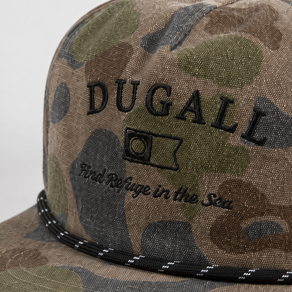 Close-up of custom black embroidered logo on a camo cotton snapback hat with rope detail across the brim.