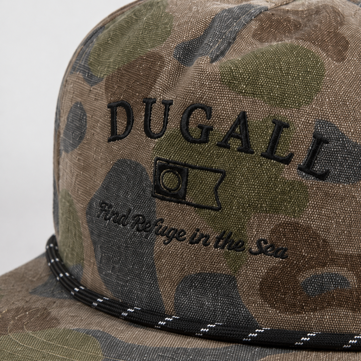 Close-up of custom black embroidered logo on a camo cotton snapback hat with rope detail across the brim.