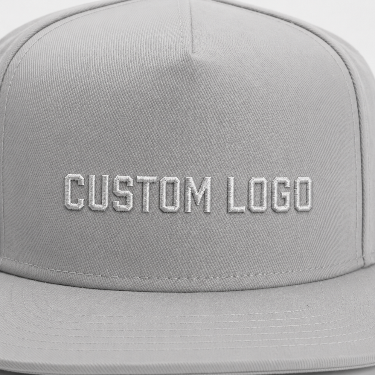 Close-up detail of embroidered “CUSTOM LOGO” text on the front panel of a light gray cotton snapback hat