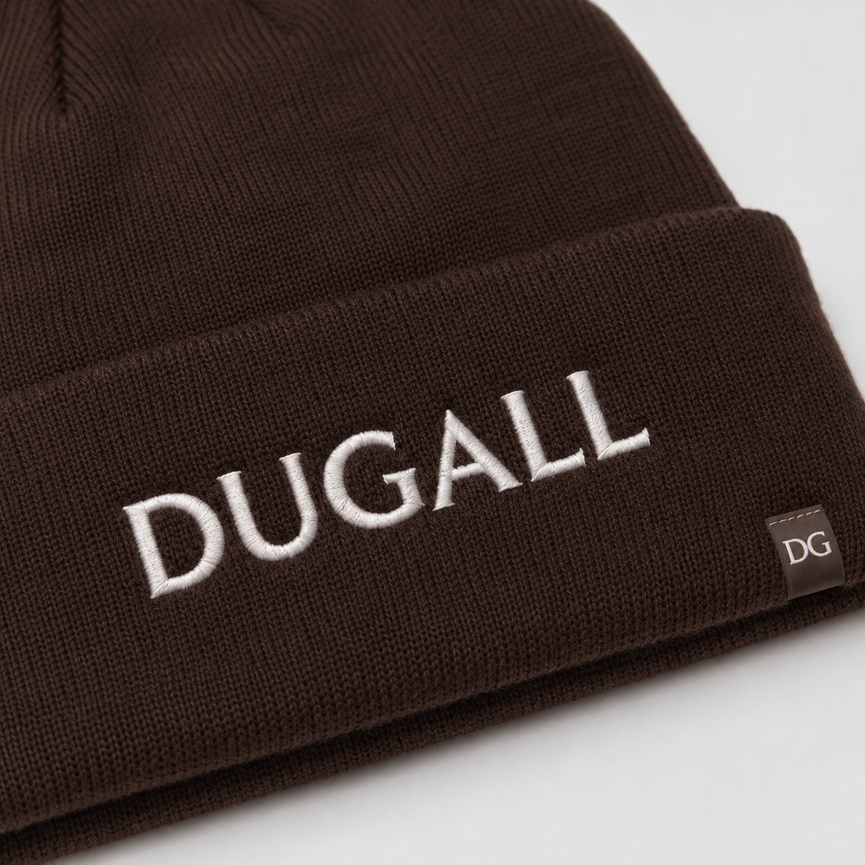 Close-up of white embroidered DUGALL logo and side label on a brown acrylic spandex beanie.